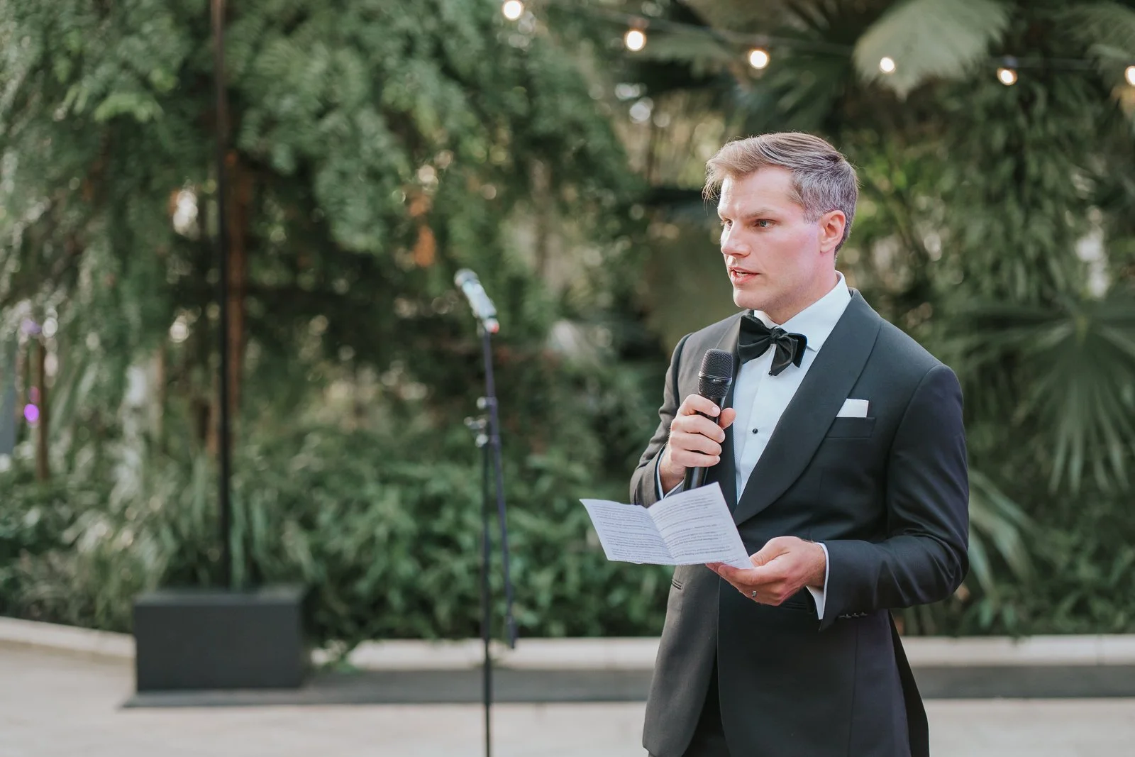  Groom giving a heartfelt speech during a wedding reception in the Temperate House at Kew Gardens, framed by tropical plants and soft evening light. 
