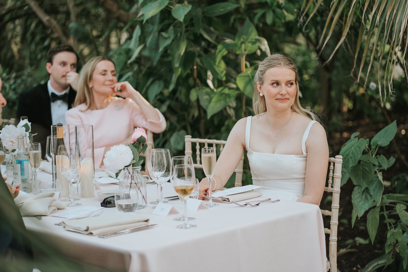  Bride listening to wedding speeches during her reception in the Temperate House at Kew Gardens, surrounded by lush greenery and elegant floral décor. 
