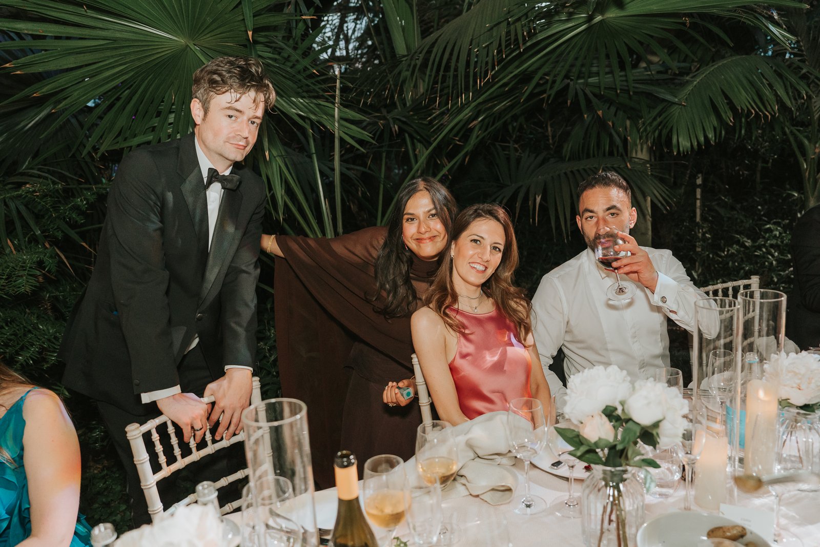  Wedding guests gathered around a reception table in the Temperate House at Kew Gardens, smiling and enjoying speeches surrounded by tropical foliage and elegant candlelit décor. 