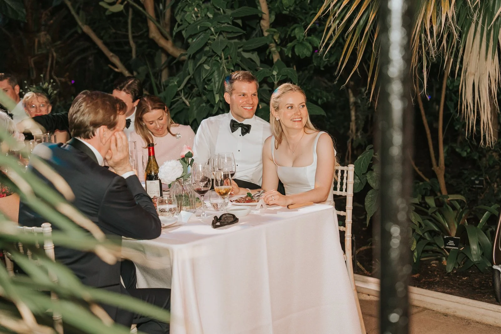  Bride, groom and guests listening attentively to speeches during a wedding reception in the Temperate House at Kew Gardens, framed by exotic palms and botanical surroundings. 