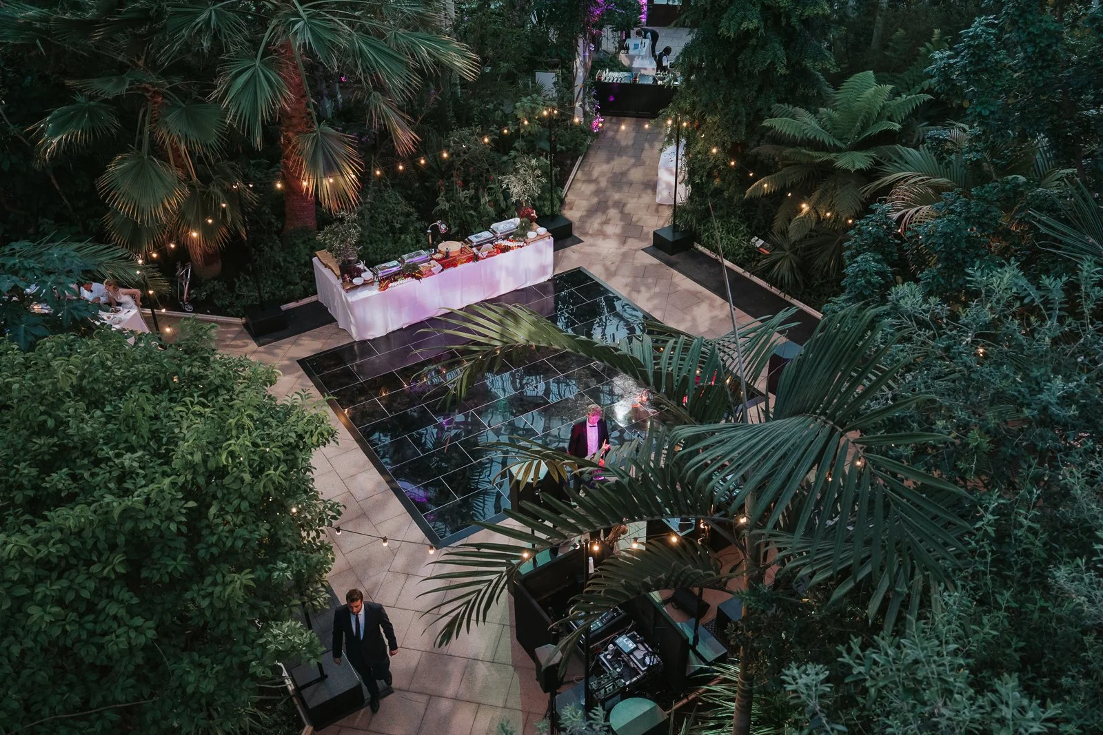  Atmospheric evening view of the wedding reception in the Temperate House at Kew Gardens, with guests dancing beneath colourful lights surrounded by tropical plants. 