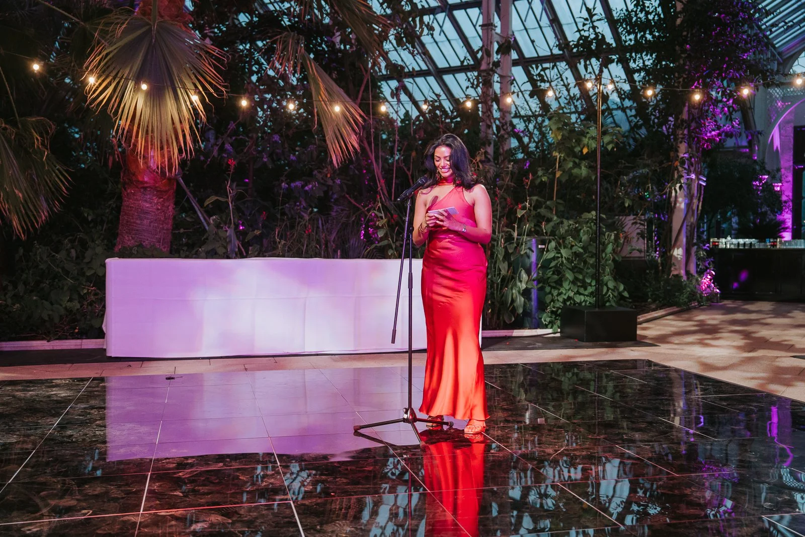  Wedding guest delivering a heartfelt speech on the dance floor of the Temperate House at Kew Gardens, illuminated by colourful evening lighting and surrounded by tropical plants. 