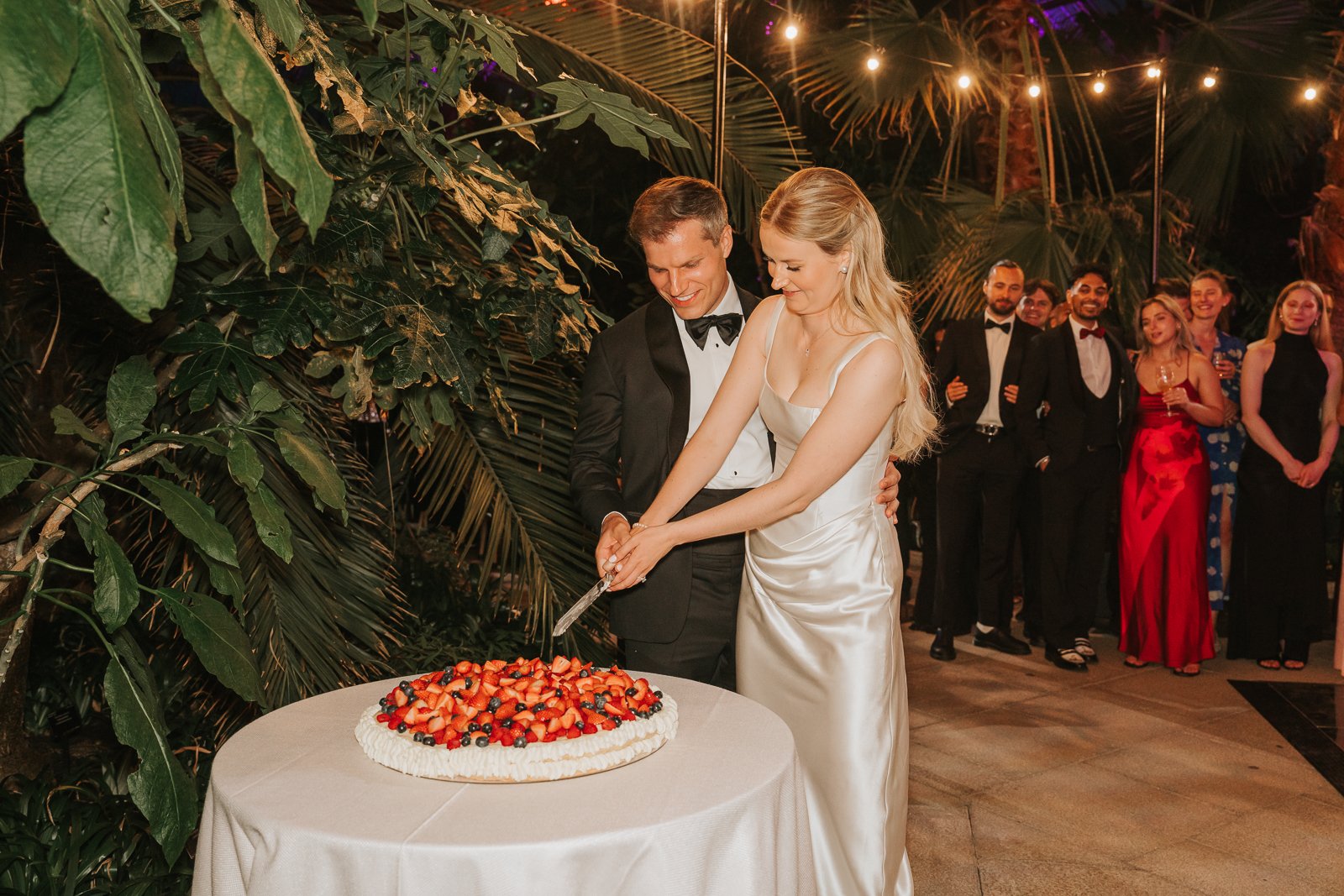  Bride and groom cutting a fruit-topped wedding cake in the Temperate House at Kew Gardens, with guests gathered around beneath warm festoon lighting. 