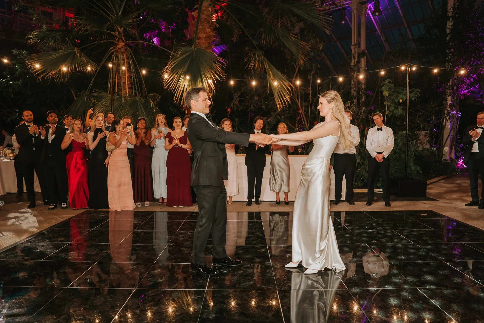 Bride and groom sharing their first dance in the Temperate House at Kew Gardens, surrounded by family and friends beneath tropical palms and twinkling lights. 