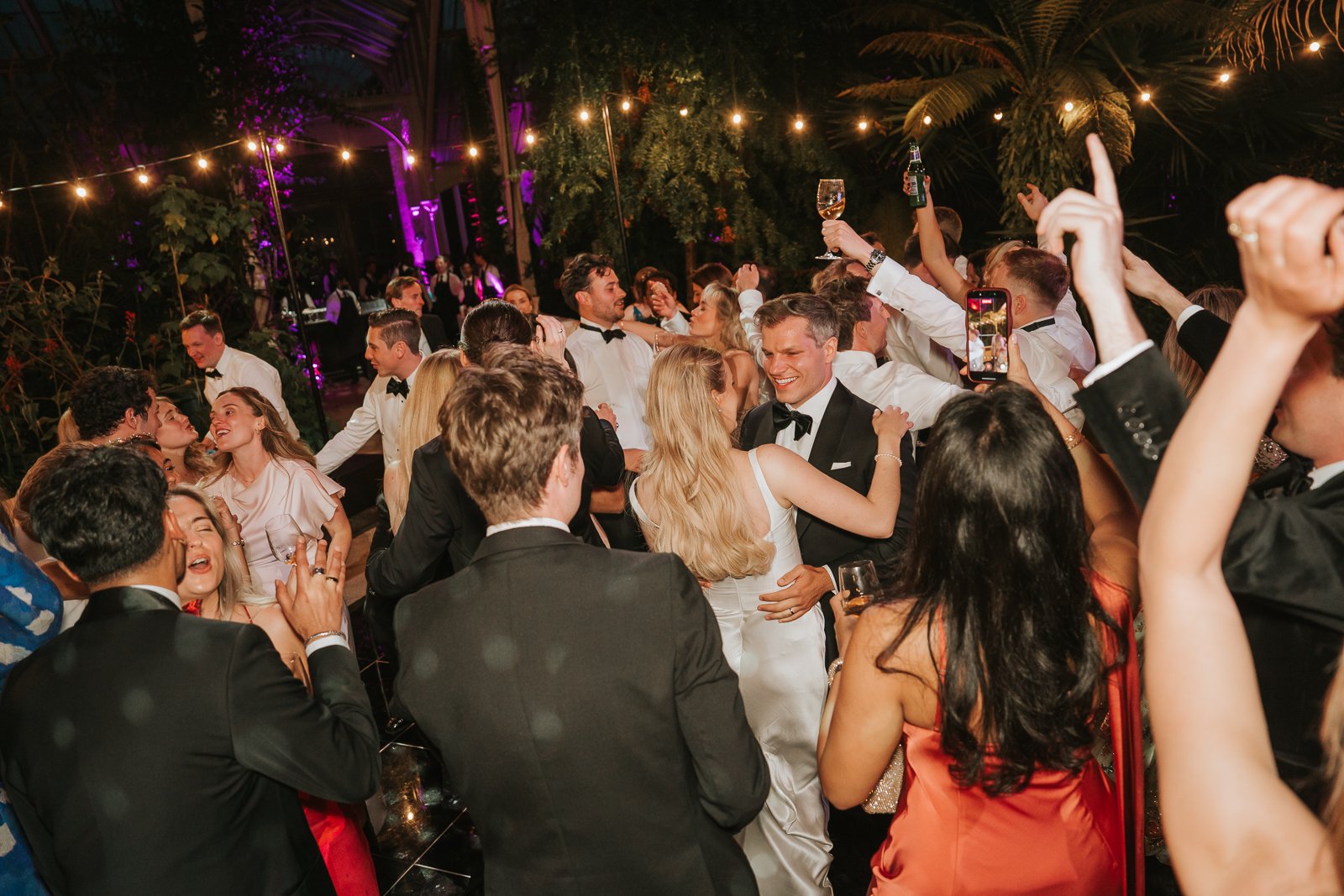  Guests enjoying the dance floor during a lively wedding reception in the Temperate House at Kew Gardens, captured in a natural documentary style. 