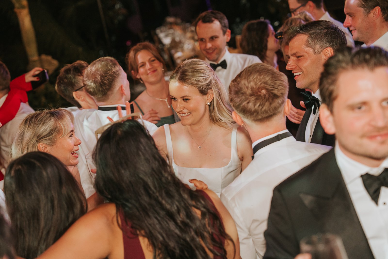  Bride dancing with friends and family at a wedding reception in the Temperate House at Kew Gardens, surrounded by joyful guests and tropical plants. 