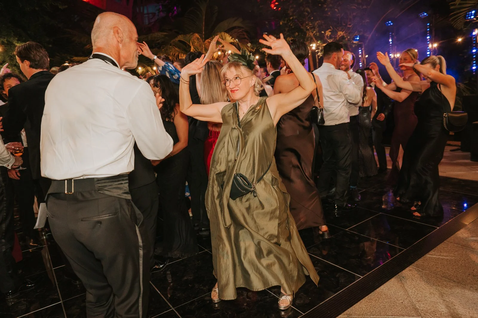  Wedding guest dancing enthusiastically on the dance floor in the Temperate House at Kew Gardens, with colourful lighting and a lively party atmosphere. 