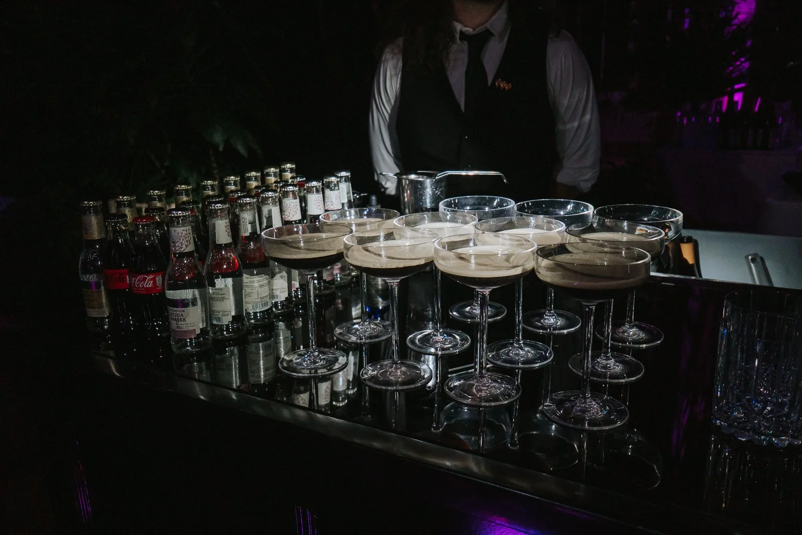  Espresso martinis prepared for wedding guests at the Temperate House at Kew Gardens, displayed on a sleek bar beneath atmospheric purple lighting. 