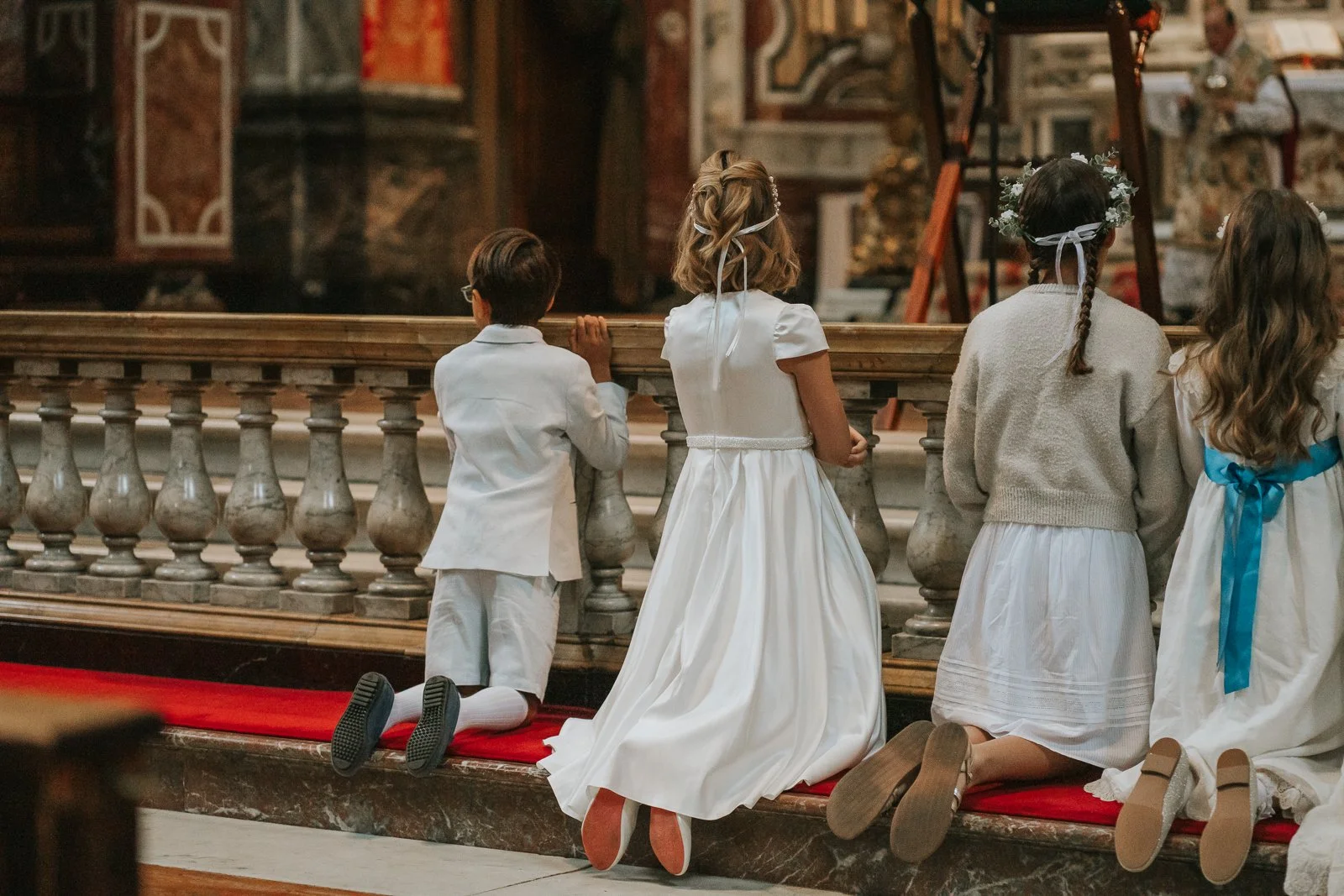  Children dressed in white ceremonial outfits kneeling at the altar rail inside Brompton Oratory, London, during a First Holy Communion service surrounded by the church’s magnificent marble interior. 