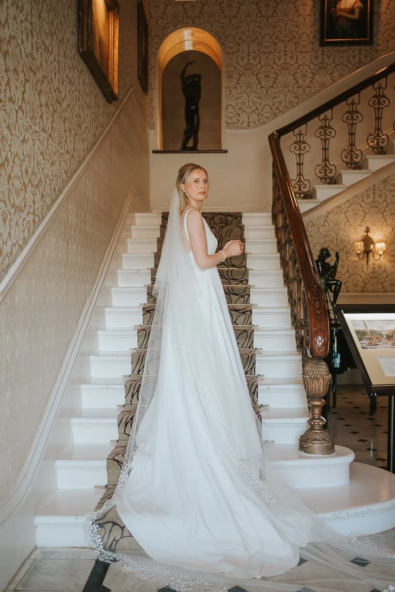  Bride in a flowing white wedding gown and cathedral-length veil posing on the elegant staircase at The Petersham Hotel in Richmond, surrounded by ornate Victorian décor and warm natural light, creating a timeless and romantic wedding portrait. 