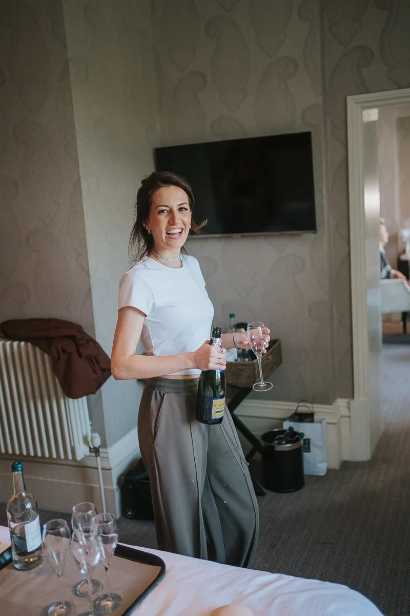  Bridesmaid opening a bottle of champagne in a luxury room at The Petersham Hotel, Richmond, during relaxed wedding morning preparations. 