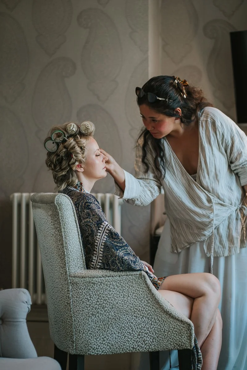  Make-up artist applying the finishing touches to the bride's make-up while she sits in a hotel room at The Petersham Hotel on her wedding morning. 