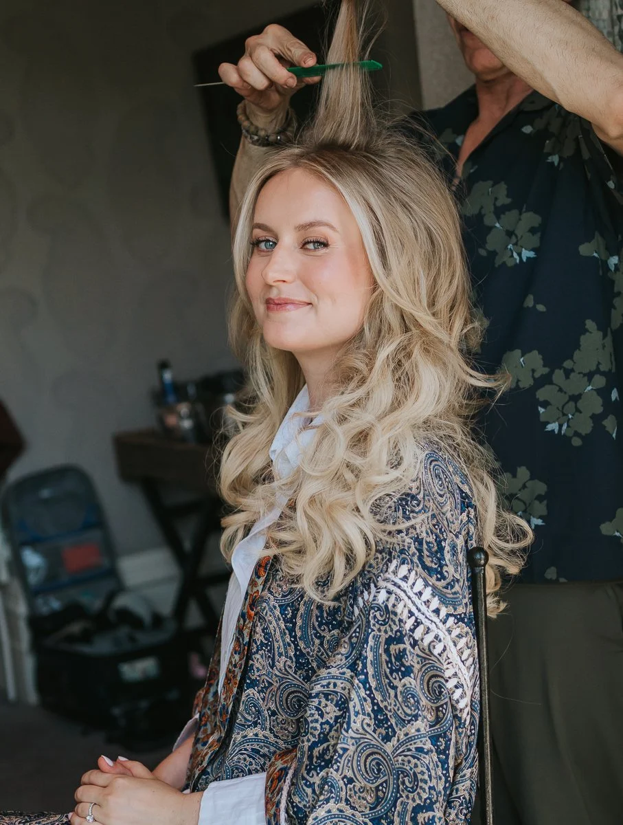  Bride smiling as her hairstylist adds volume and finishing touches to her blonde curls during bridal preparations at The Petersham Hotel. 