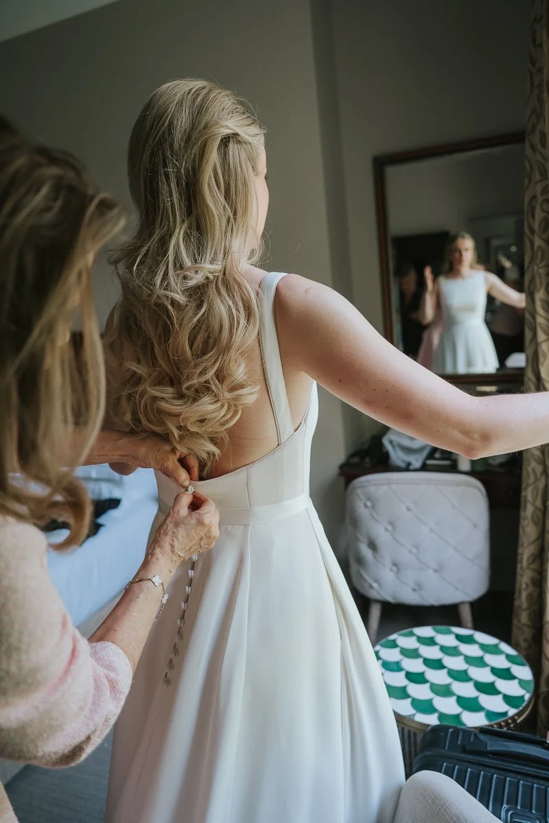  Bride's mother fastening the button-back wedding dress as the bride stands by the window in her hotel suite at The Petersham Hotel. 