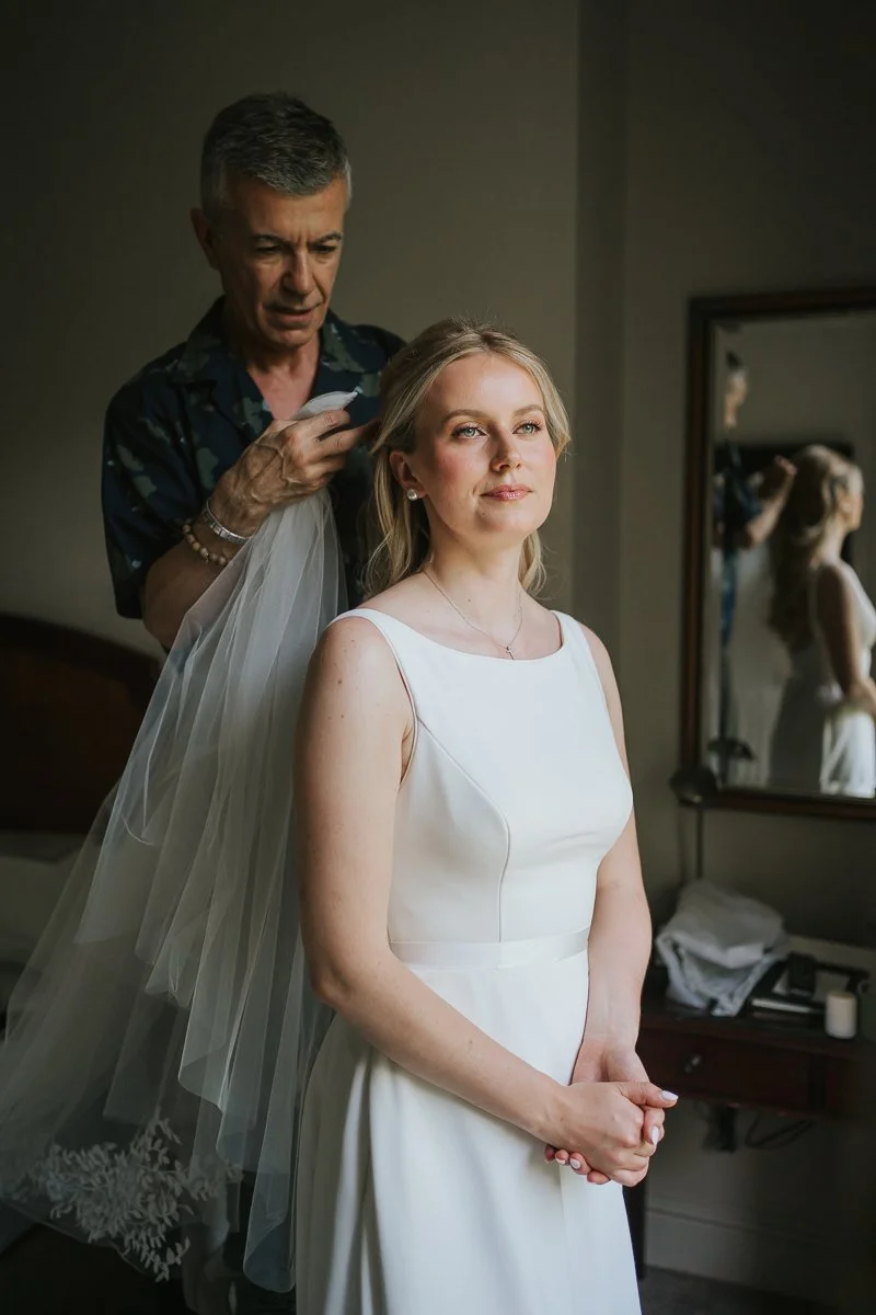  Bride standing patiently while her hairstylist secures her wedding veil, capturing a quiet moment before the ceremony at The Petersham Hotel. 