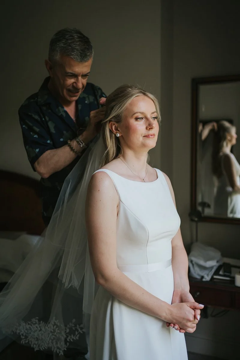  Bride in a simple white wedding gown as her veil is carefully positioned by her hairstylist in a luxury room at The Petersham Hotel, Richmond. 