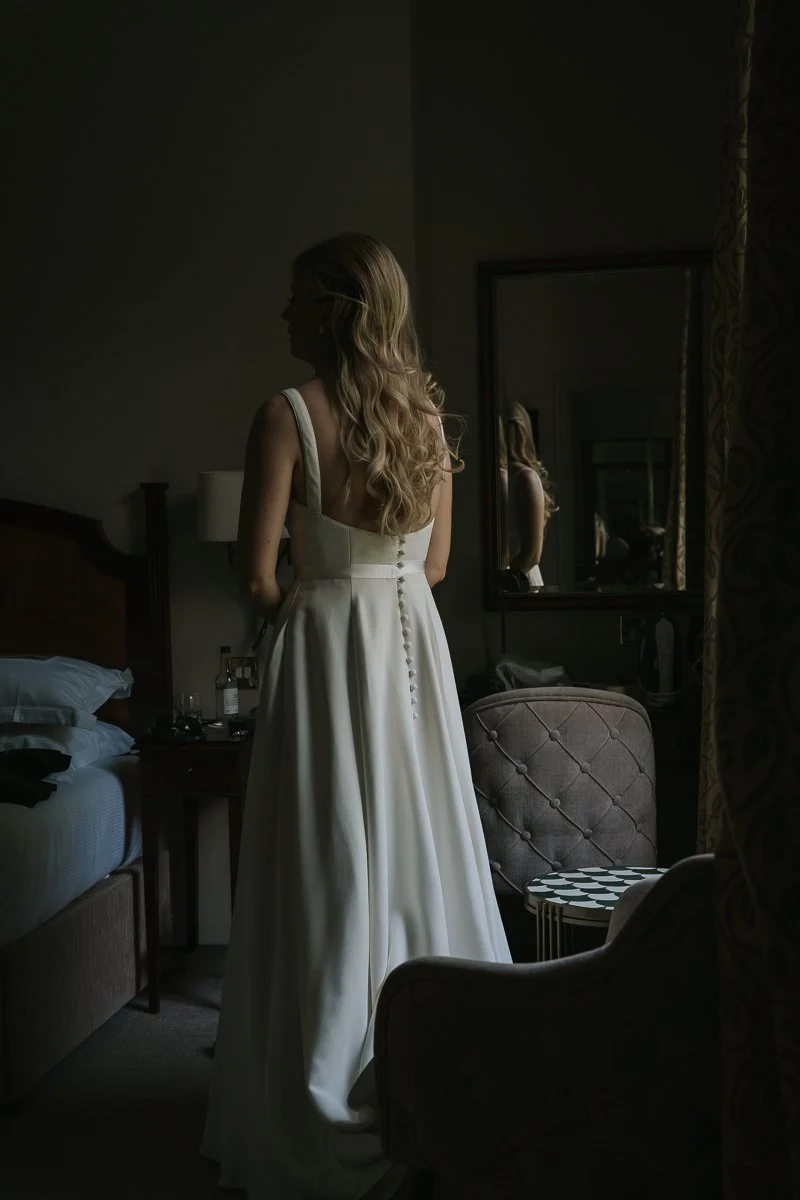  Atmospheric bridal portrait showing the back of a bride's wedding dress with elegant button detailing, photographed in soft window light at The Petersham Hotel. 
