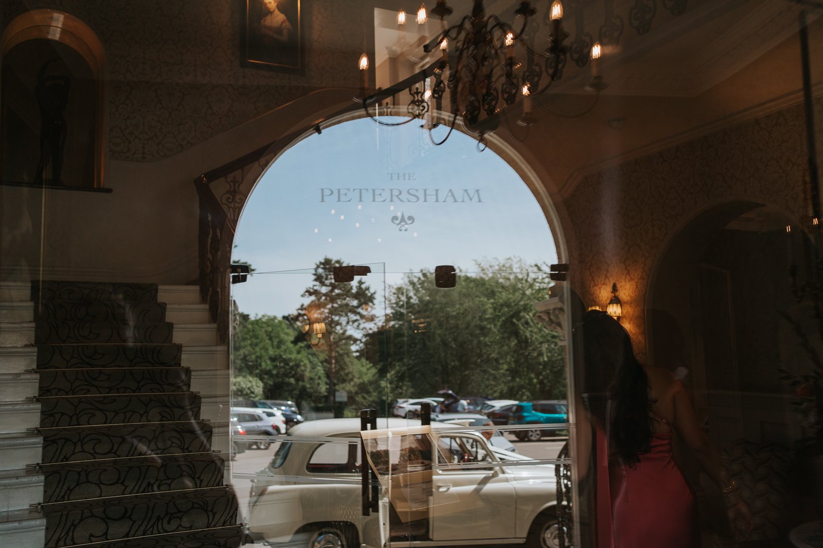  Creative reflection photograph inside The Petersham Hotel showing the grand staircase, chandelier and vintage wedding taxi arriving outside the hotel entrance in Richmond. 