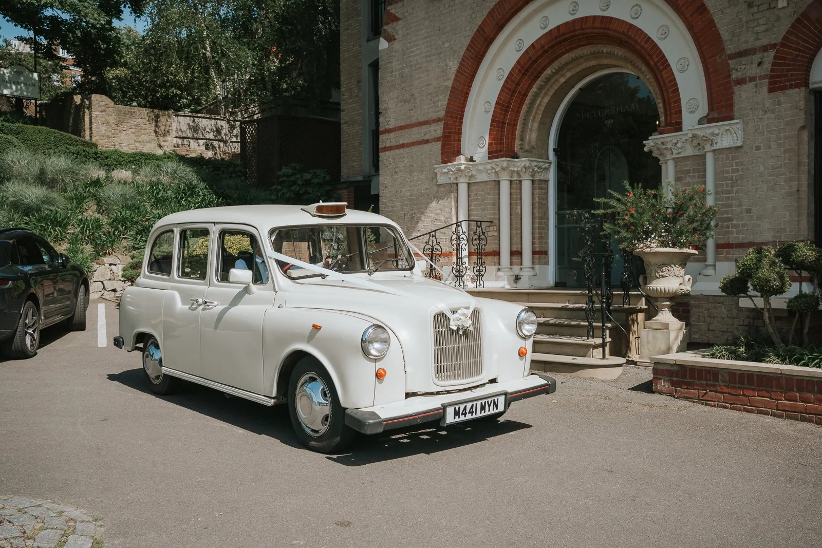  Vintage white London taxi decorated for a wedding parked outside The Petersham Hotel in Richmond, with the hotel's historic red-brick entrance creating a beautiful wedding venue backdrop. 