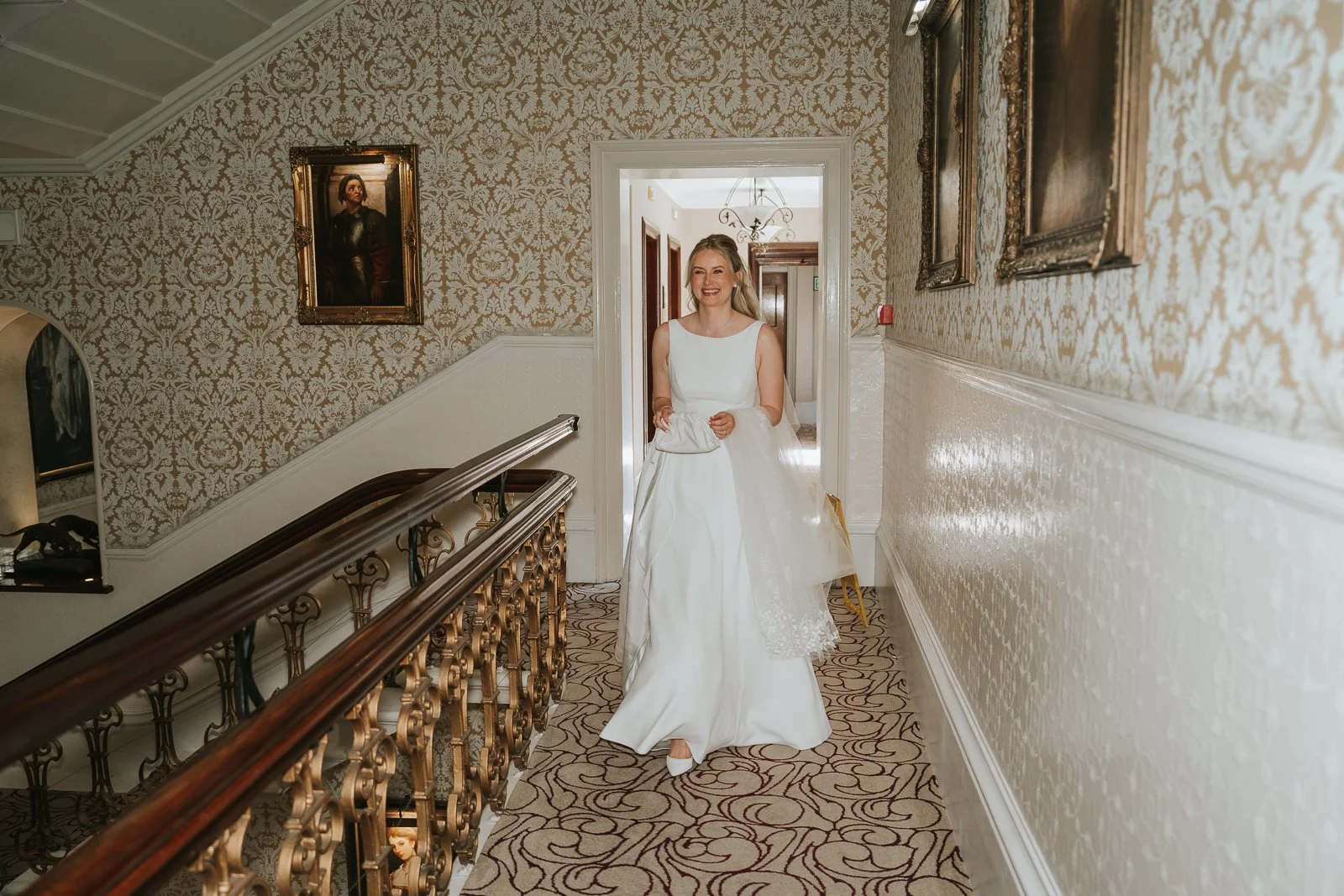  Bride walking through the elegant upper hallway at The Petersham Hotel in Richmond, smiling as she carries her veil before the wedding ceremony, surrounded by ornate wallpaper and classic Victorian details. 