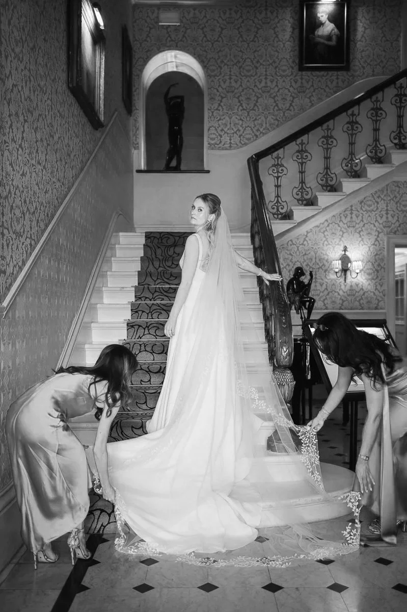  Bride standing on the grand staircase at The Petersham Hotel in Richmond while two bridesmaids in pink satin dresses carefully arrange her wedding gown and lace-edged veil, creating an elegant behind-the-scenes bridal preparation moment. 