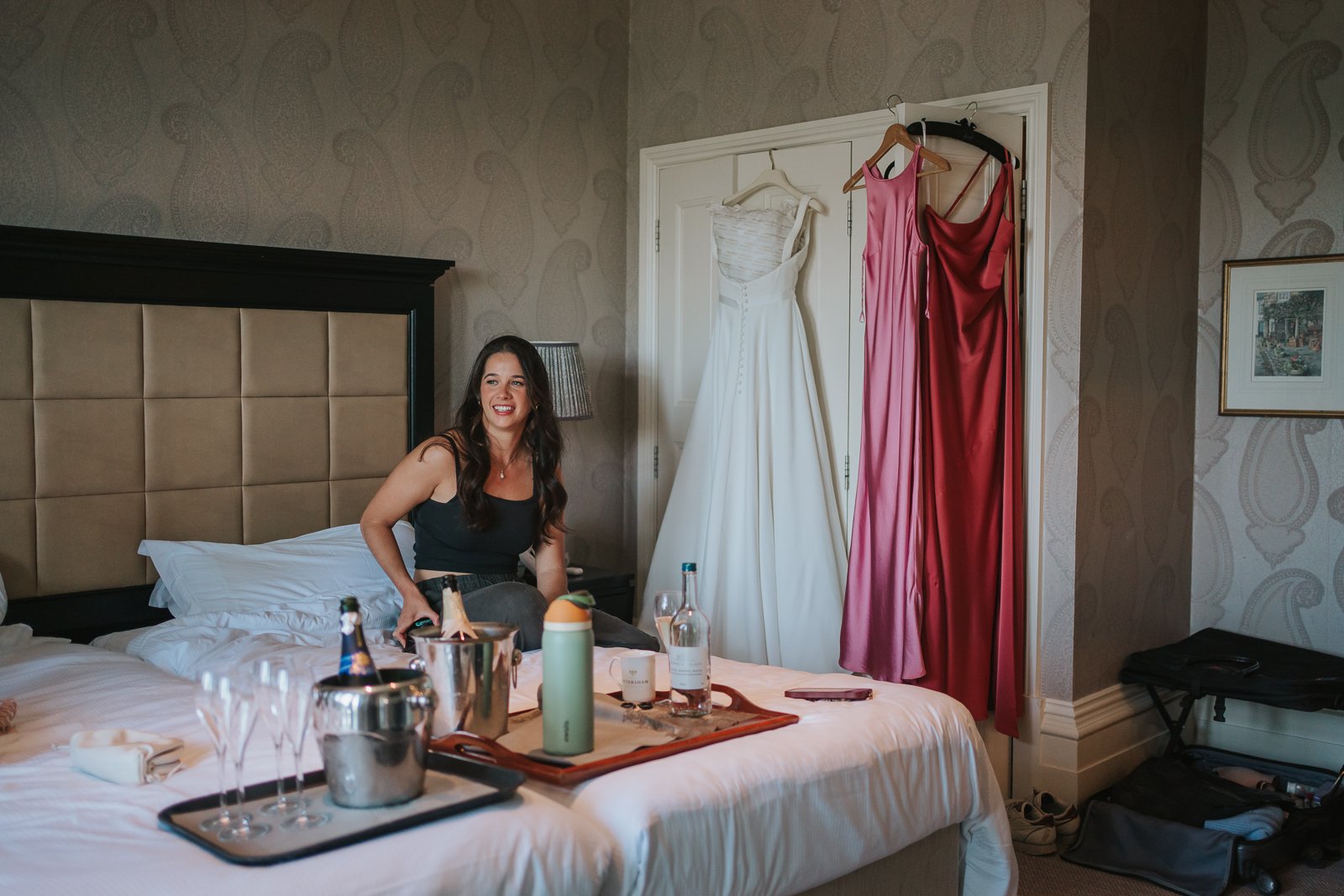  Bridesmaid relaxing in a hotel room surrounded by wedding details, champagne and hanging dresses during bridal preparations at The Petersham Hotel. 