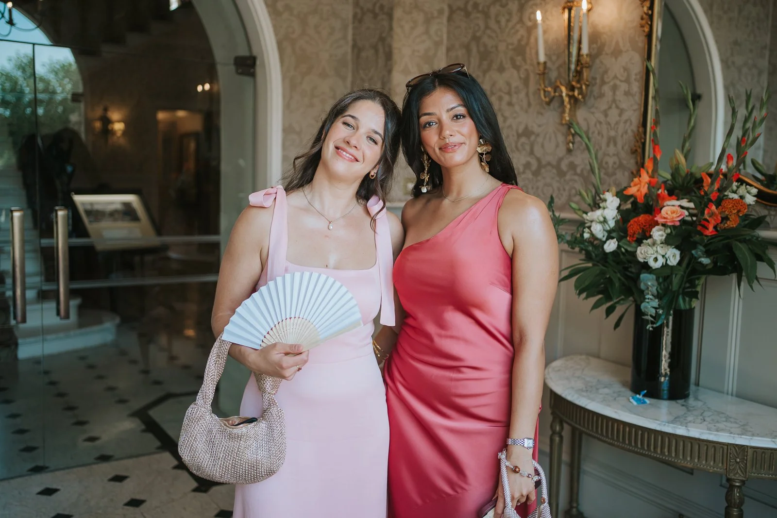  Two bridesmaids in elegant pink dresses posing in the grand entrance hall of The Petersham Hotel before the wedding ceremony. 