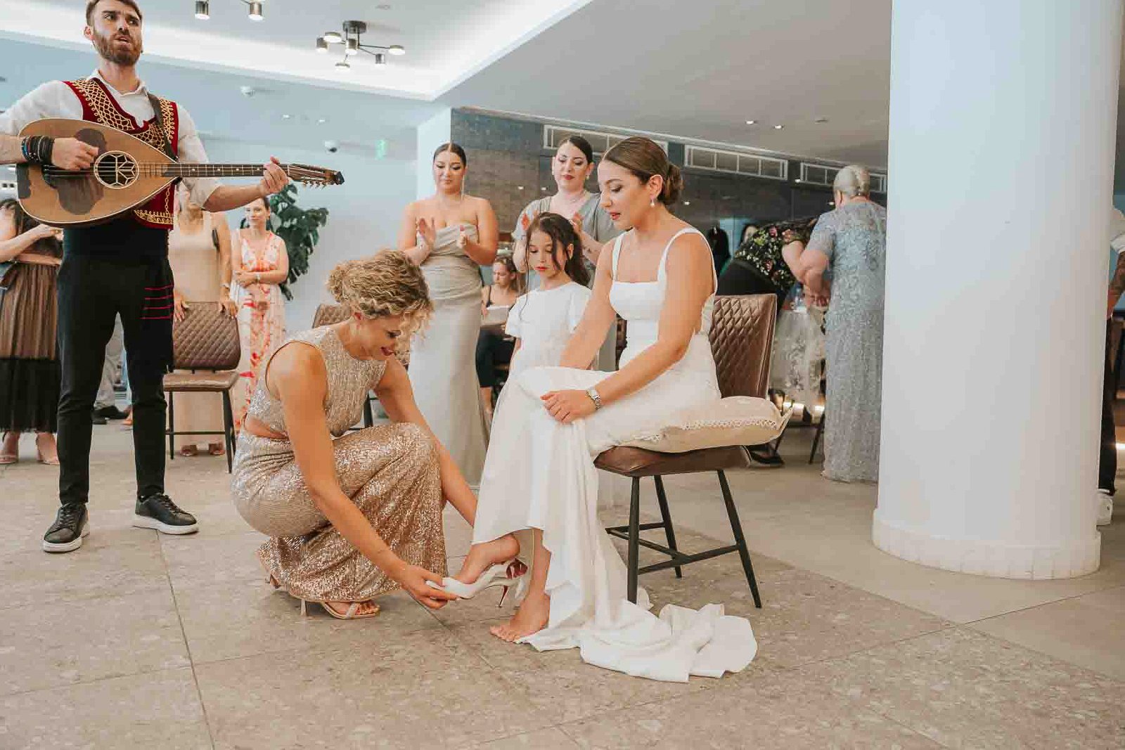  Bride seated as a family member helps her put on a shoe during a traditional Greek wedding reception ritual, with live music and guests gathered around. 