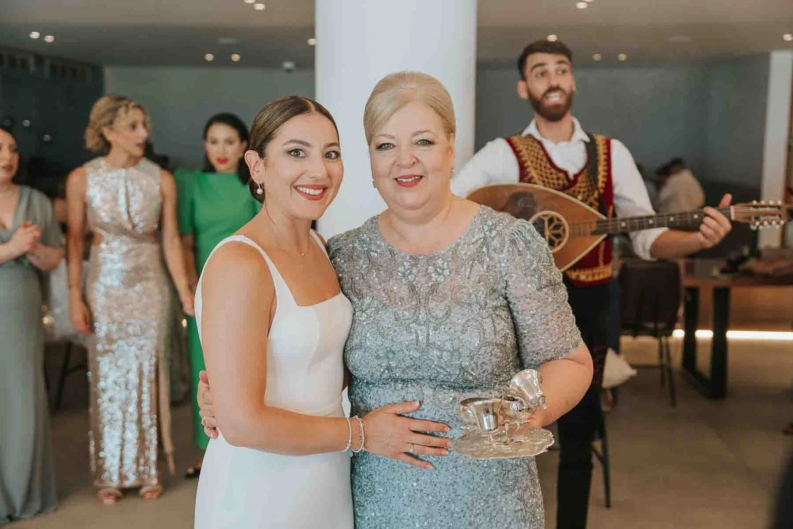 Bride posing with her mother during a Greek wedding celebration, surrounded by family, guests and traditional Greek musicians. 