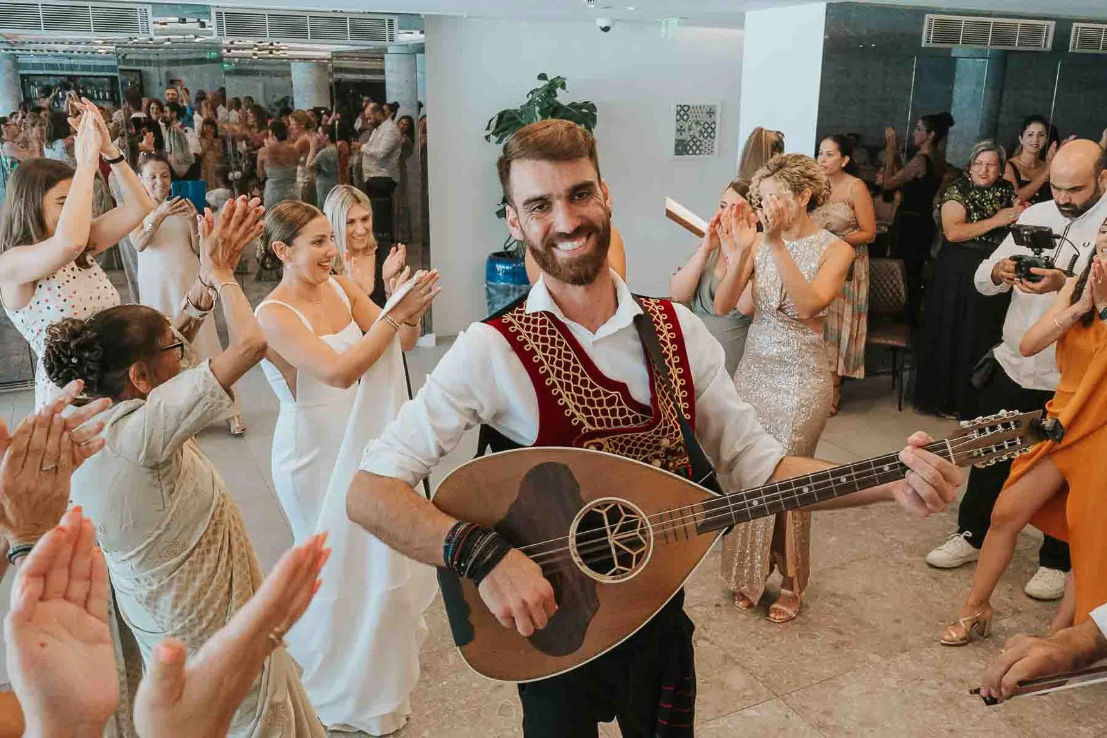  Traditional Greek wedding musician playing the bouzouki while guests clap, dance and celebrate during a lively wedding reception. 