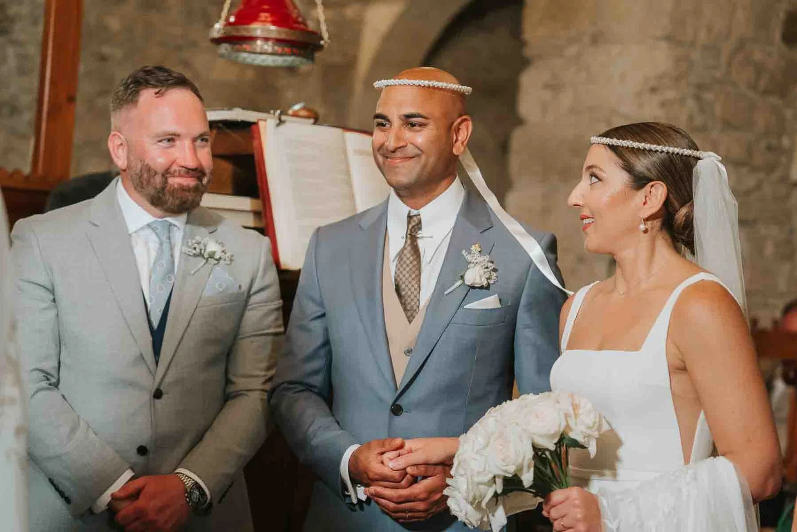  Bride and groom wearing their stefana crowns during the sacrament of marriage, sharing a joyful moment with their koumbaro. 