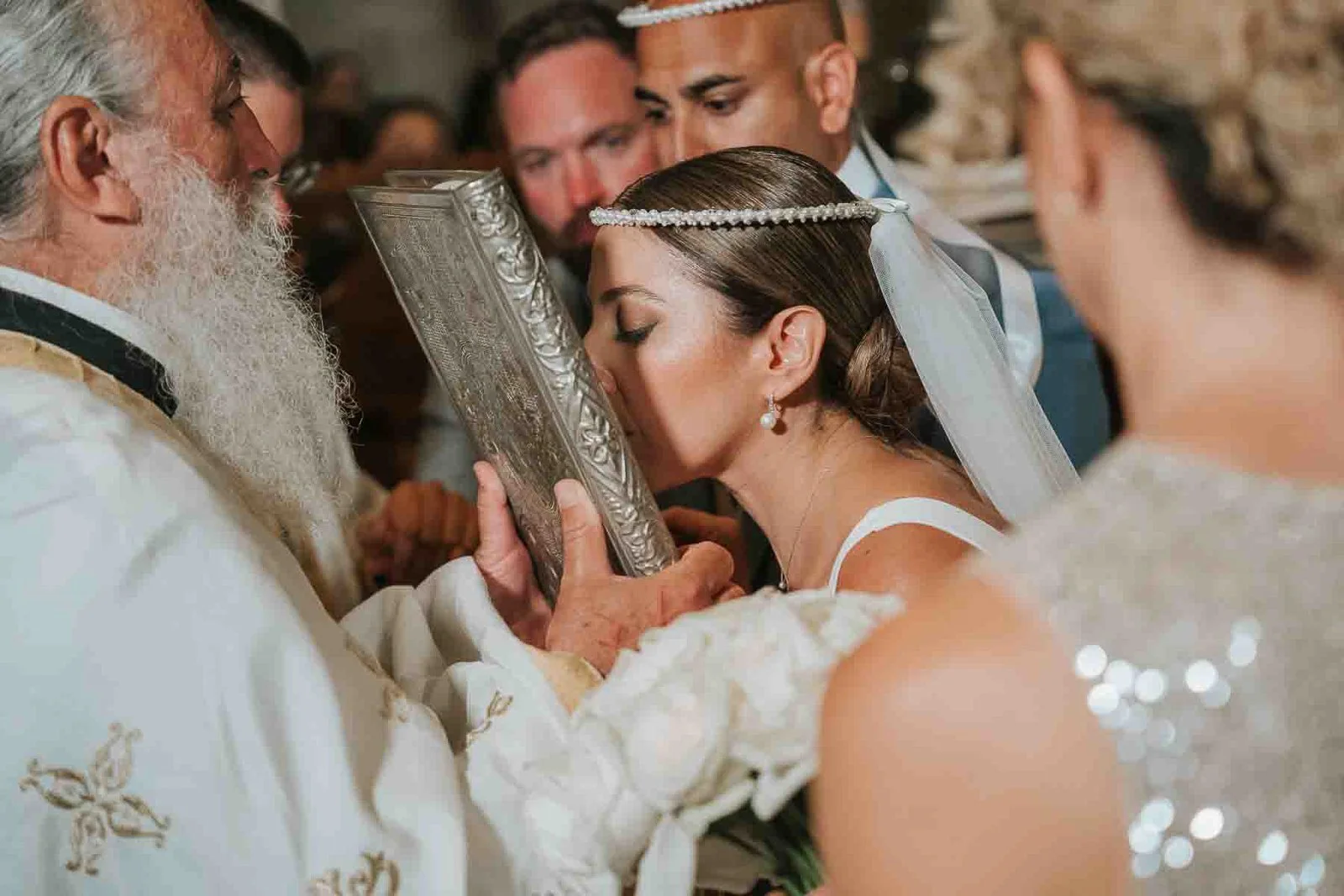  Bride kissing a sacred icon during a Greek Orthodox wedding ceremony, surrounded by family and wedding guests. 