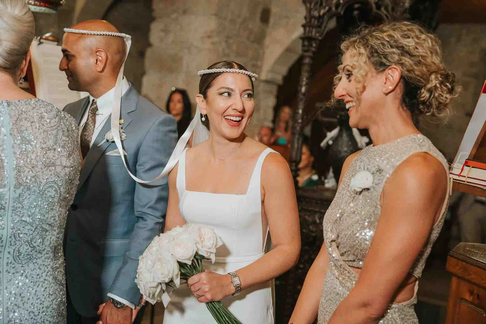  Bride smiling at her koumbara during the Greek Orthodox wedding ceremony, wearing traditional stefana crowns inside the historic church. 