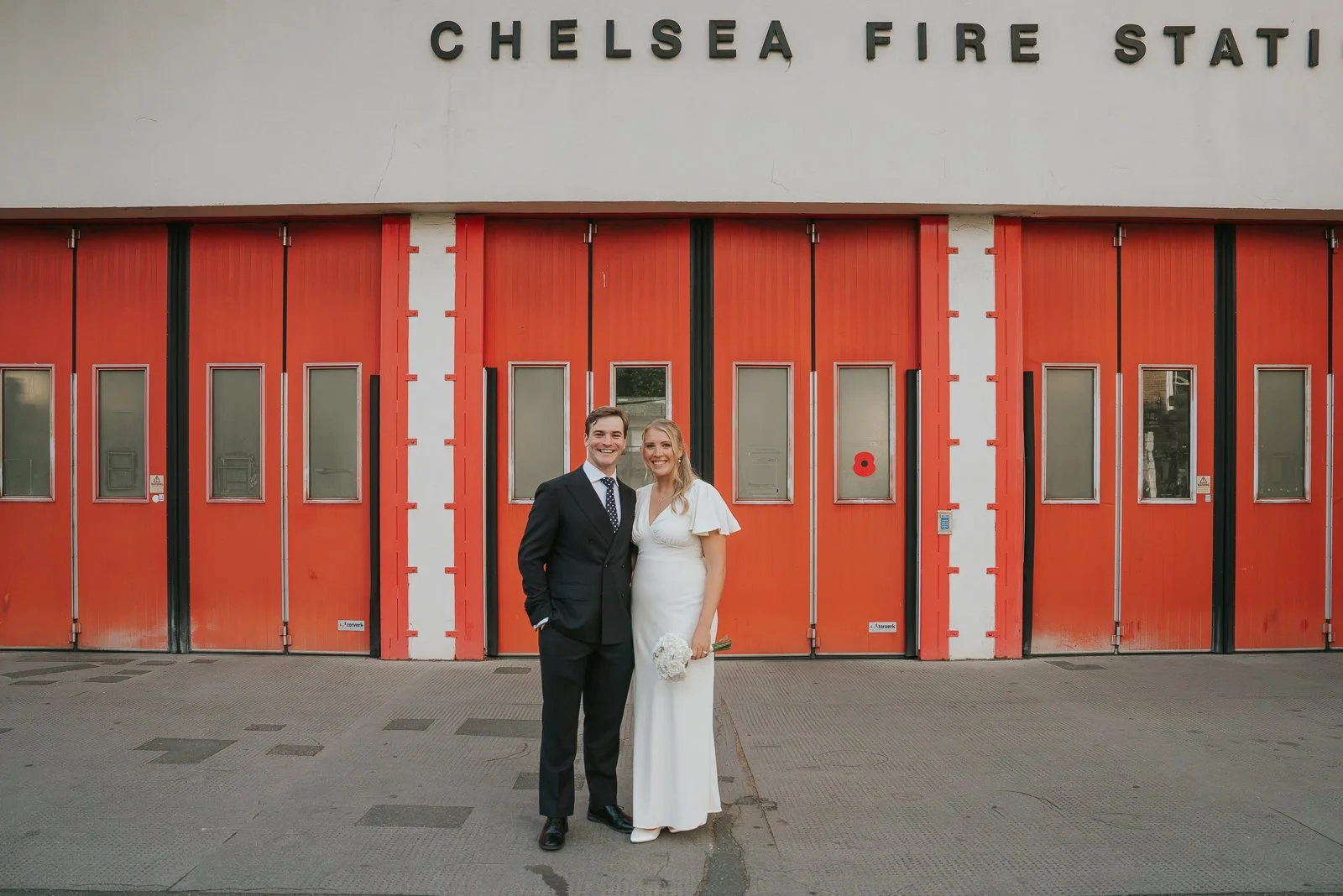  Newly married couple posing outside Chelsea Fire Station in London, with its striking red doors providing a bold and contemporary backdrop for stylish Chelsea wedding photography. 