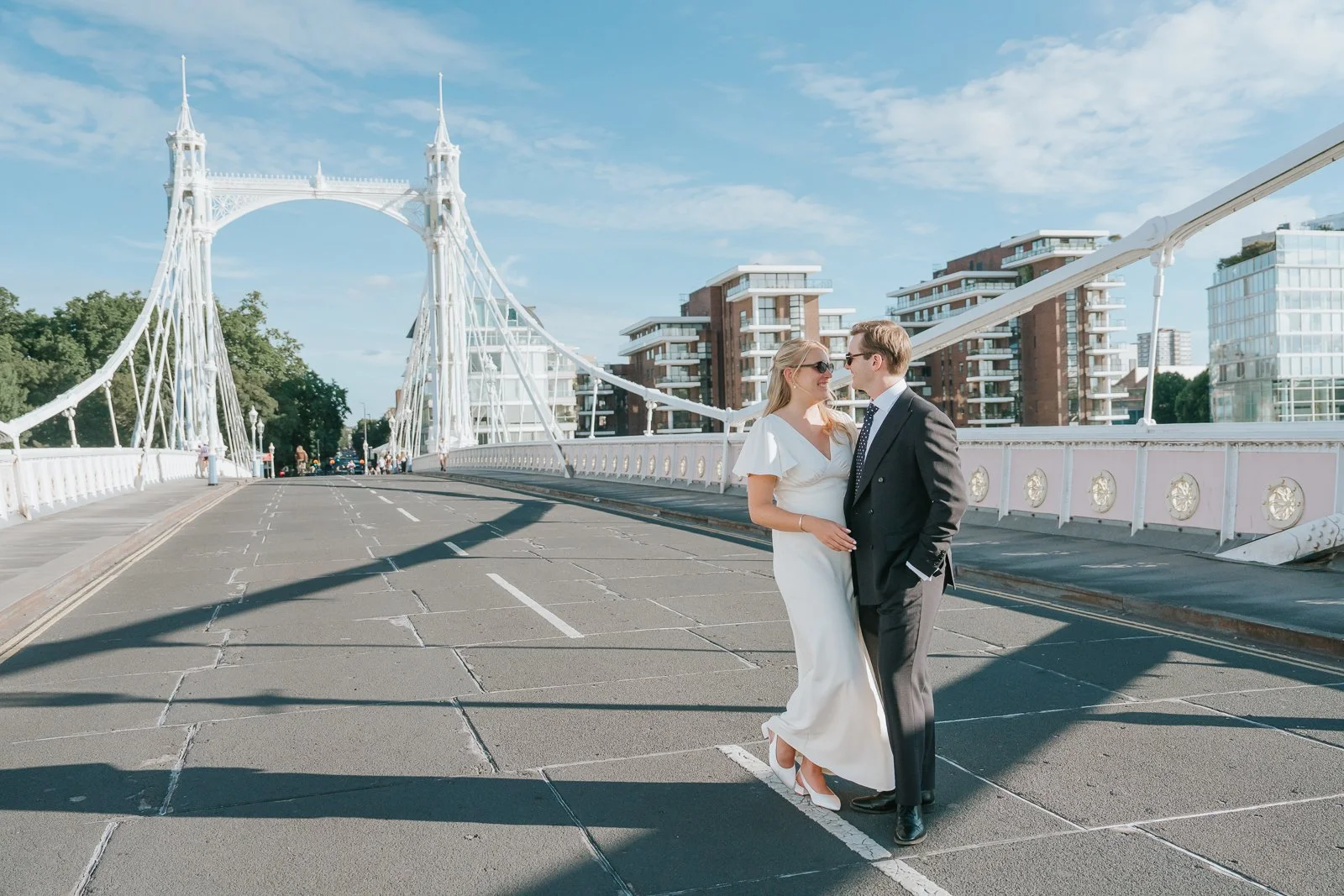  Newly married couple sharing a quiet moment on Albert Bridge in Chelsea, London, with the iconic white suspension bridge and bright summer sky creating a romantic wedding portrait setting. 