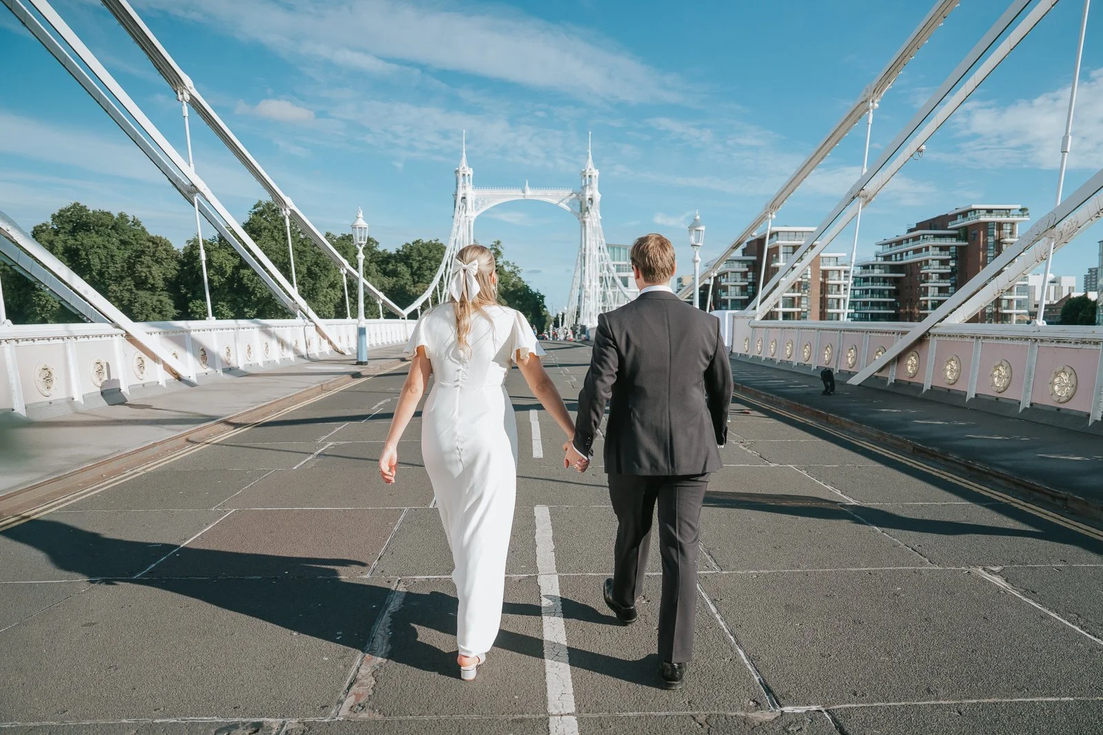  Bride and groom walking hand in hand across Albert Bridge in Chelsea, London, enjoying a quiet moment together beneath the bridge's elegant Victorian architecture on a sunny day. 