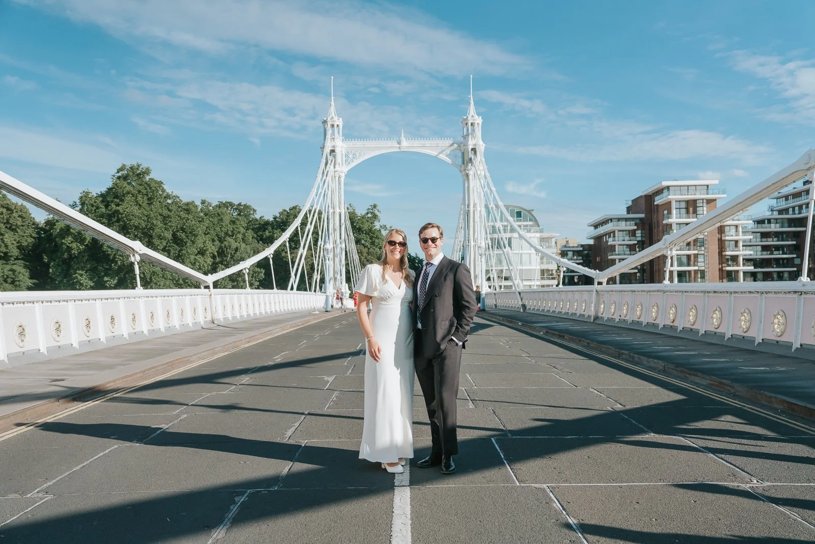  Bride and groom wearing sunglasses and posing in the centre of Albert Bridge in Chelsea, with the iconic white suspension bridge and blue summer sky creating a timeless London wedding portrait. 