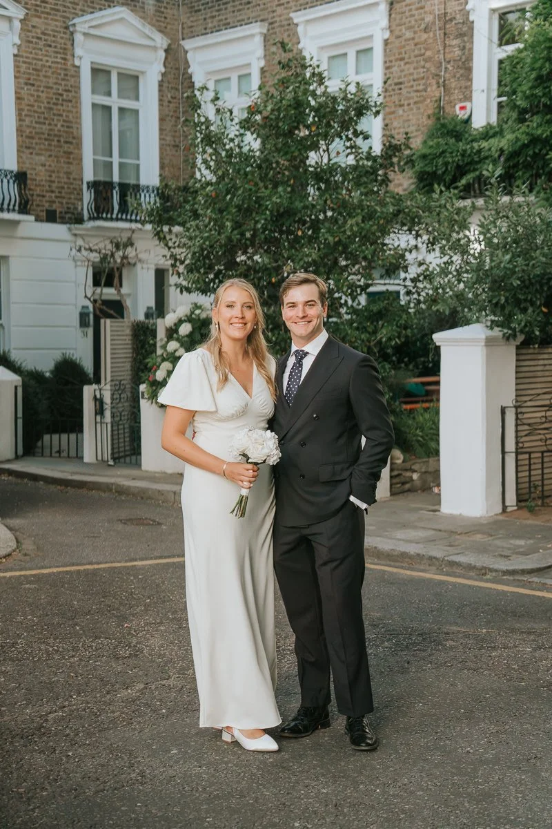  Bride and groom posing for a portrait on a picturesque Chelsea street, framed by traditional London townhouses and greenery, during an intimate city wedding photoshoot. 