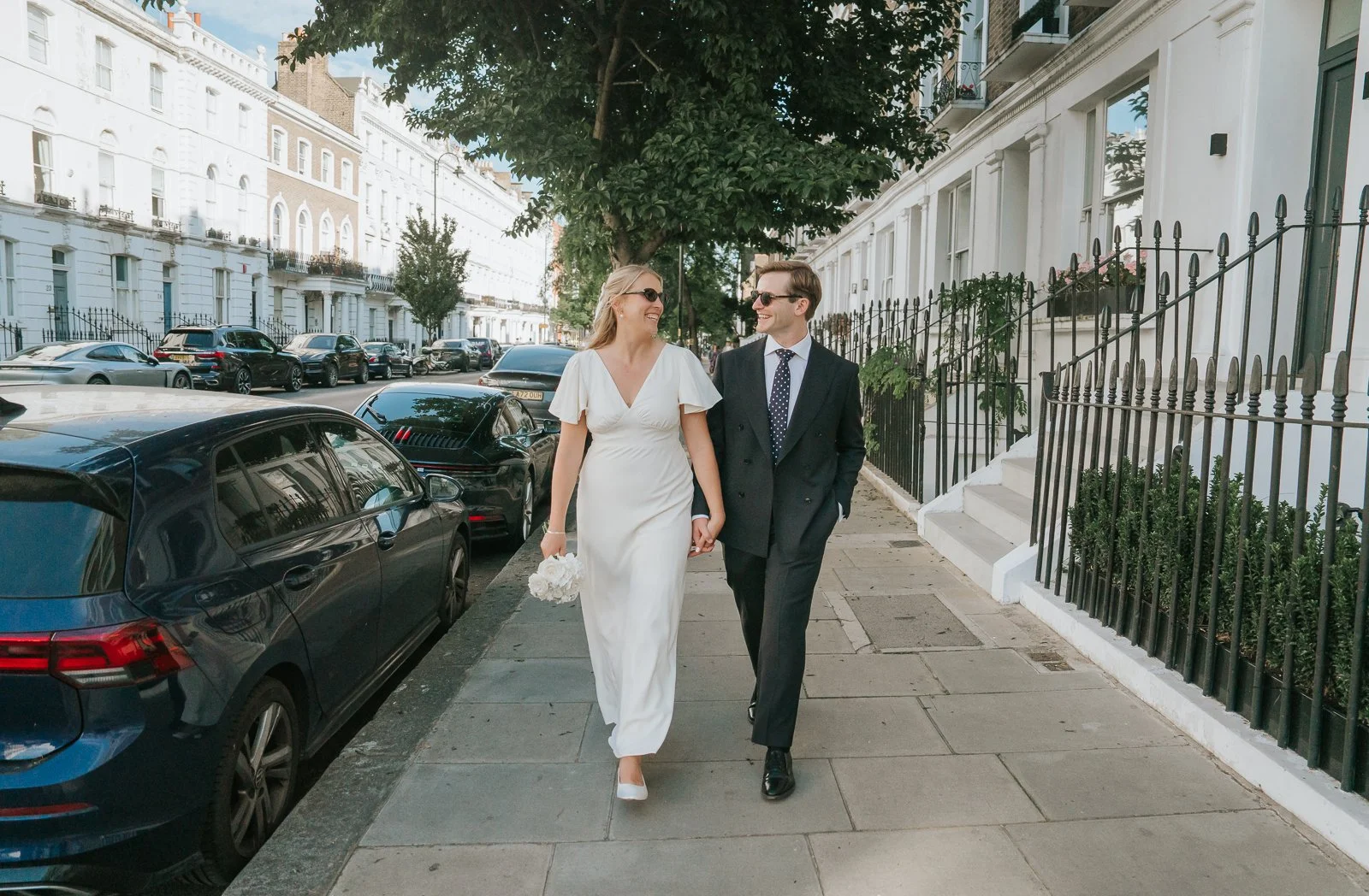   Bride and groom walking hand in hand through the elegant residential streets of Chelsea, London, surrounded by white stucco townhouses and black railings, captured in a relaxed documentary wedding photography style.  