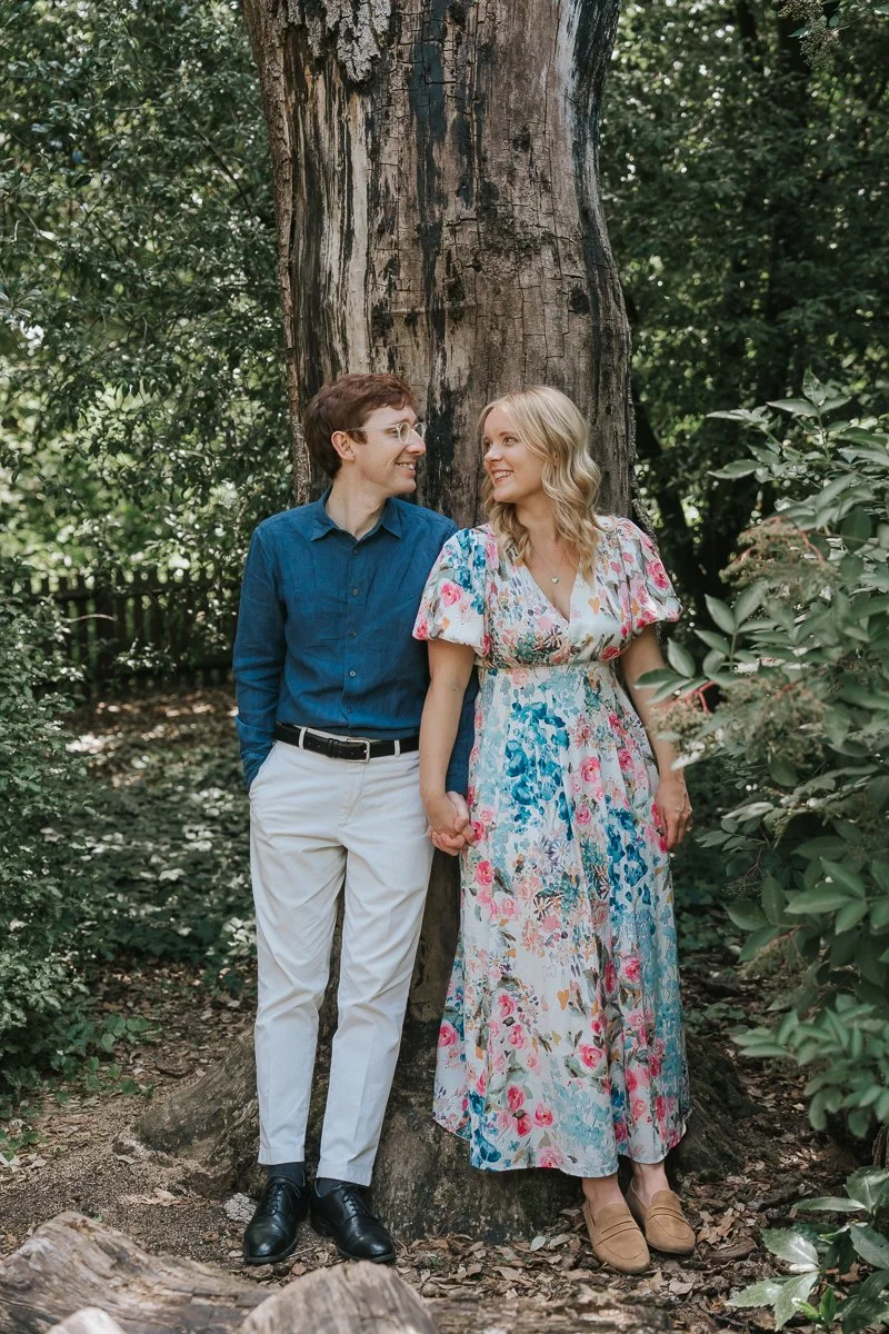 Couple standing hand in hand beneath a large tree in Battersea Park, London, sharing a smile during a relaxed summer engagement photoshoot surrounded by woodland greenery. 