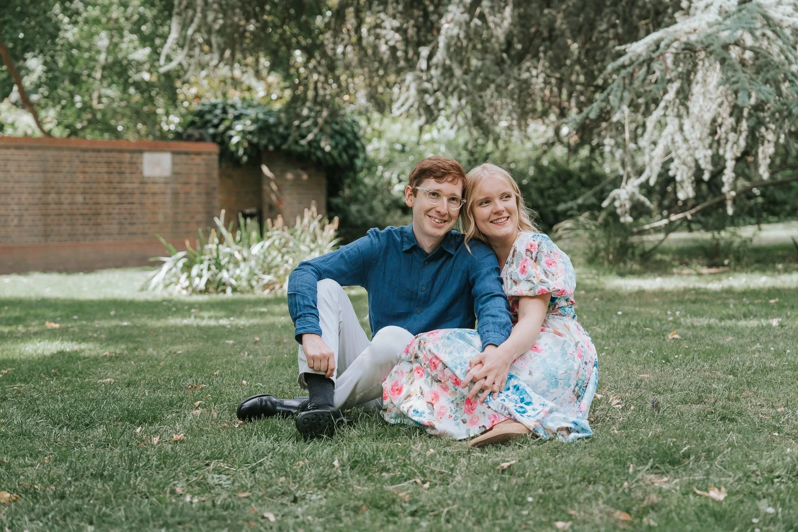  Natural engagement portrait in Battersea Park, London, featuring a couple sitting together on the grass beneath mature trees during a relaxed outdoor couples session. 
