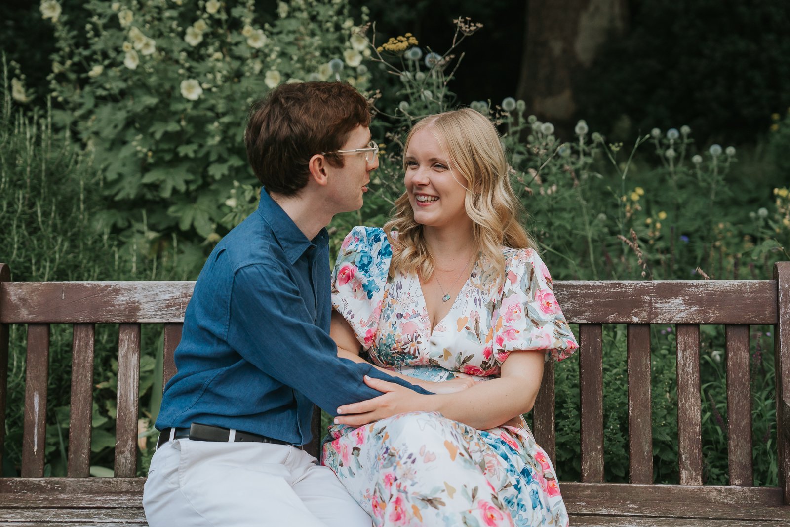  Romantic engagement photography in Battersea Park, London, with a couple sharing a conversation on a garden bench surrounded by seasonal flowers and lush greenery. 