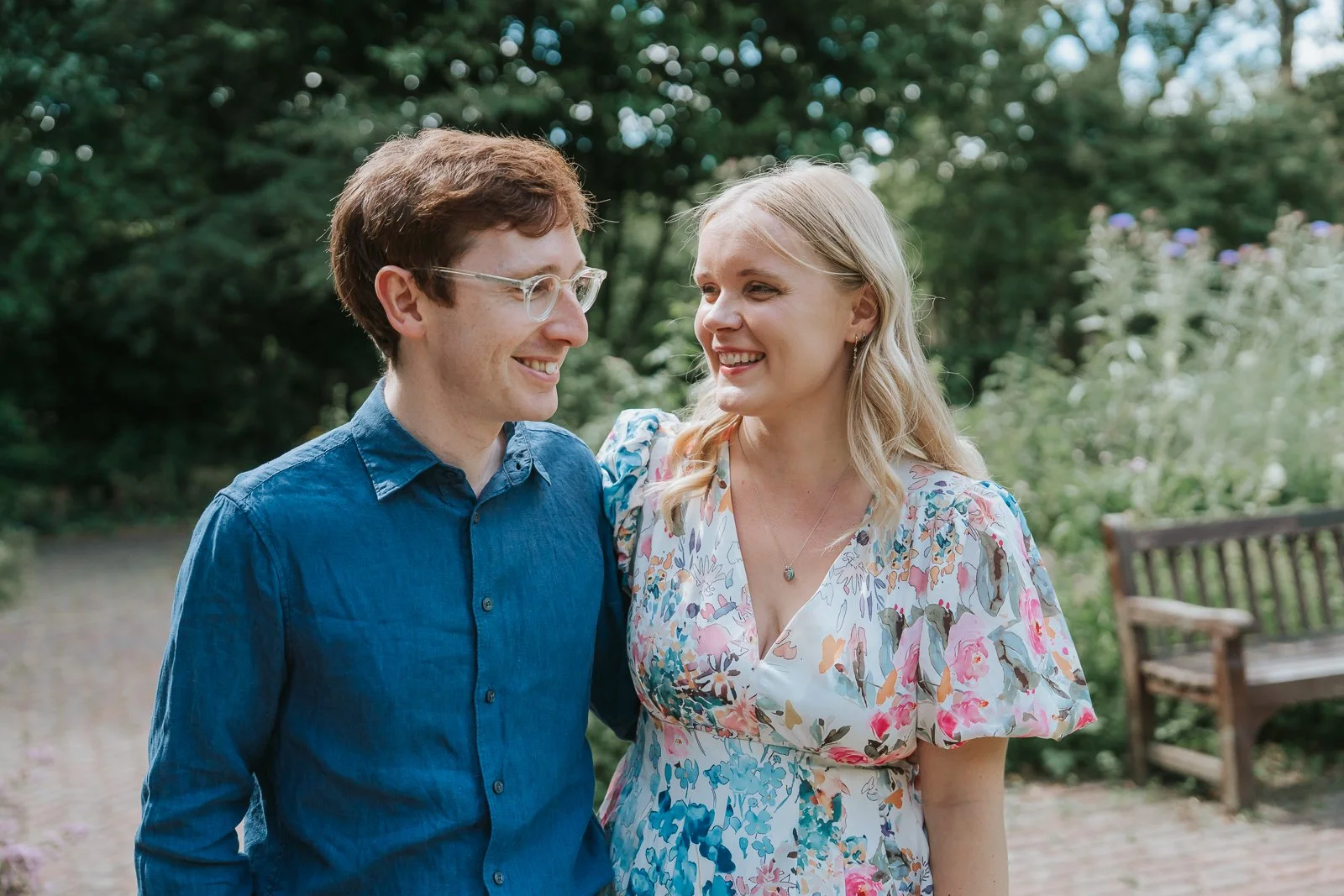  Candid engagement photography in Battersea Park, London, capturing a couple laughing together in soft summer light surrounded by flowers and greenery. 