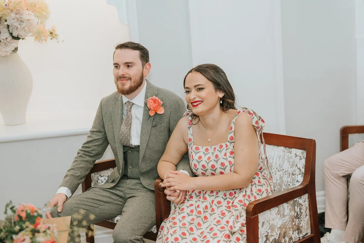   Bride and groom holding hands during their intimate wedding ceremony in the Rossetti Room at Chelsea Old Town Hall, surrounded by elegant white interiors and soft natural light.  