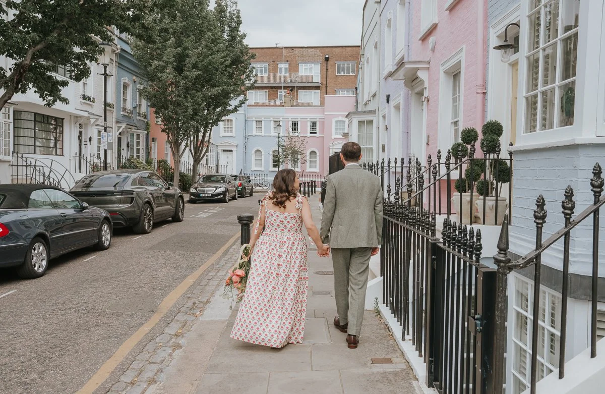   Bride and groom walking hand in hand along a colourful residential street in Chelsea, London, surrounded by pastel townhouses, during a relaxed post-ceremony wedding photoshoot.  