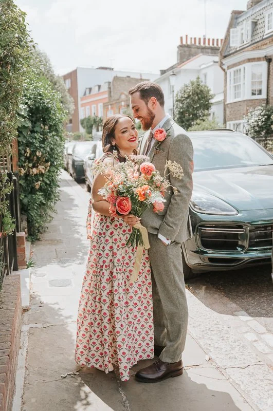  Bride and groom embracing on a quiet Chelsea street, with a vibrant bouquet and picturesque London surroundings creating a natural and intimate wedding portrait. 