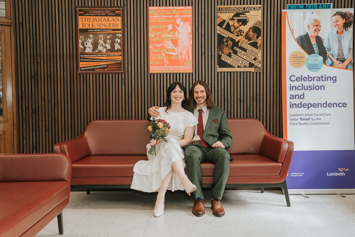  Bride and groom sitting together on a sofa inside  Lambeth Town Hall in Brixton , smiling after their intimate London registry office wedding, with colourful Lambeth heritage posters in the background. 