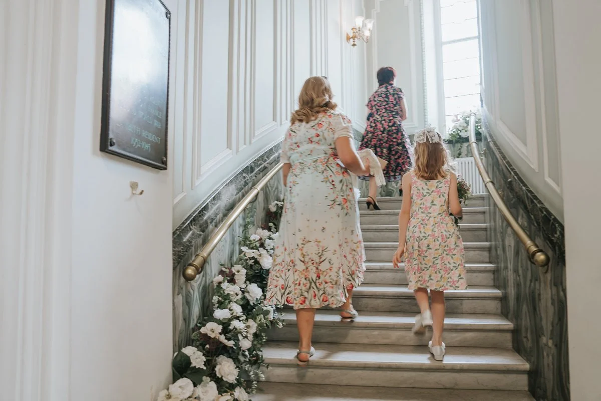  Wedding guests and a flower girl walking up the elegant marble staircase inside  Lambeth Town Hall, Brixton , capturing the anticipation before the wedding ceremony in a relaxed documentary style. 