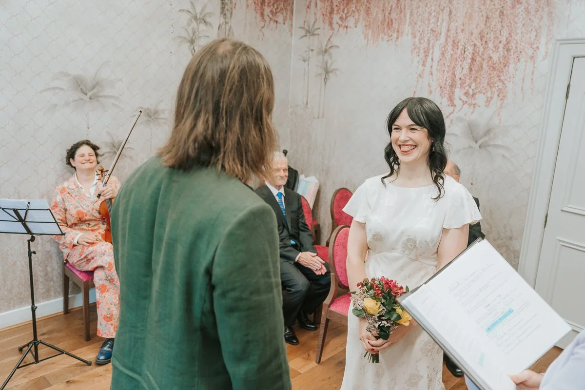  Bride and groom exchanging vows during their intimate wedding ceremony in the  Pauline Room at Lambeth Town Hall , with guests and a live violinist creating a warm, personal atmosphere. 