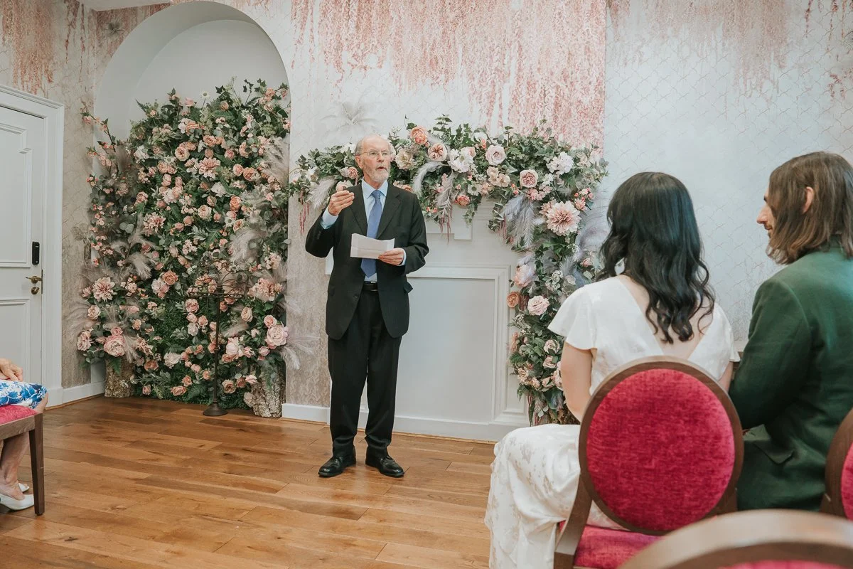  A family member giving a heartfelt reading during a civil wedding ceremony in the  Pauline Room at Lambeth Town Hall , framed by the room's elegant floral backdrop. 